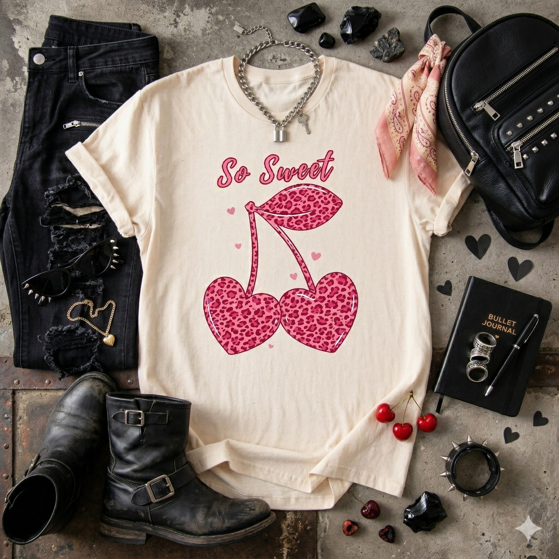 Natural t-shirt with pink leopard print cherries and 'So Sweet' text, surrounded by black boots, jeans, and accessories on a rustic surface.