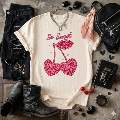 Natural t-shirt with pink leopard print cherries and 'So Sweet' text, surrounded by black boots, jeans, and accessories on a rustic surface.