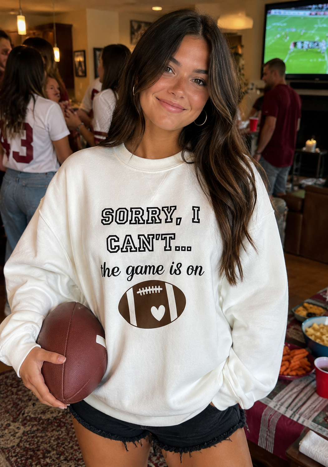 Woman wearing a sweatshirt with a football graphic and text, holding a football in a casual indoor setting.