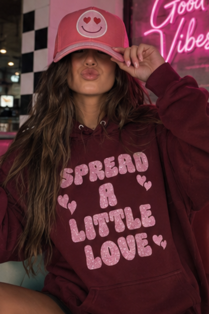 Woman wearing a maroon sweatshirt that says "Spread A Little Love" in a pink glitter graphic with hearts.