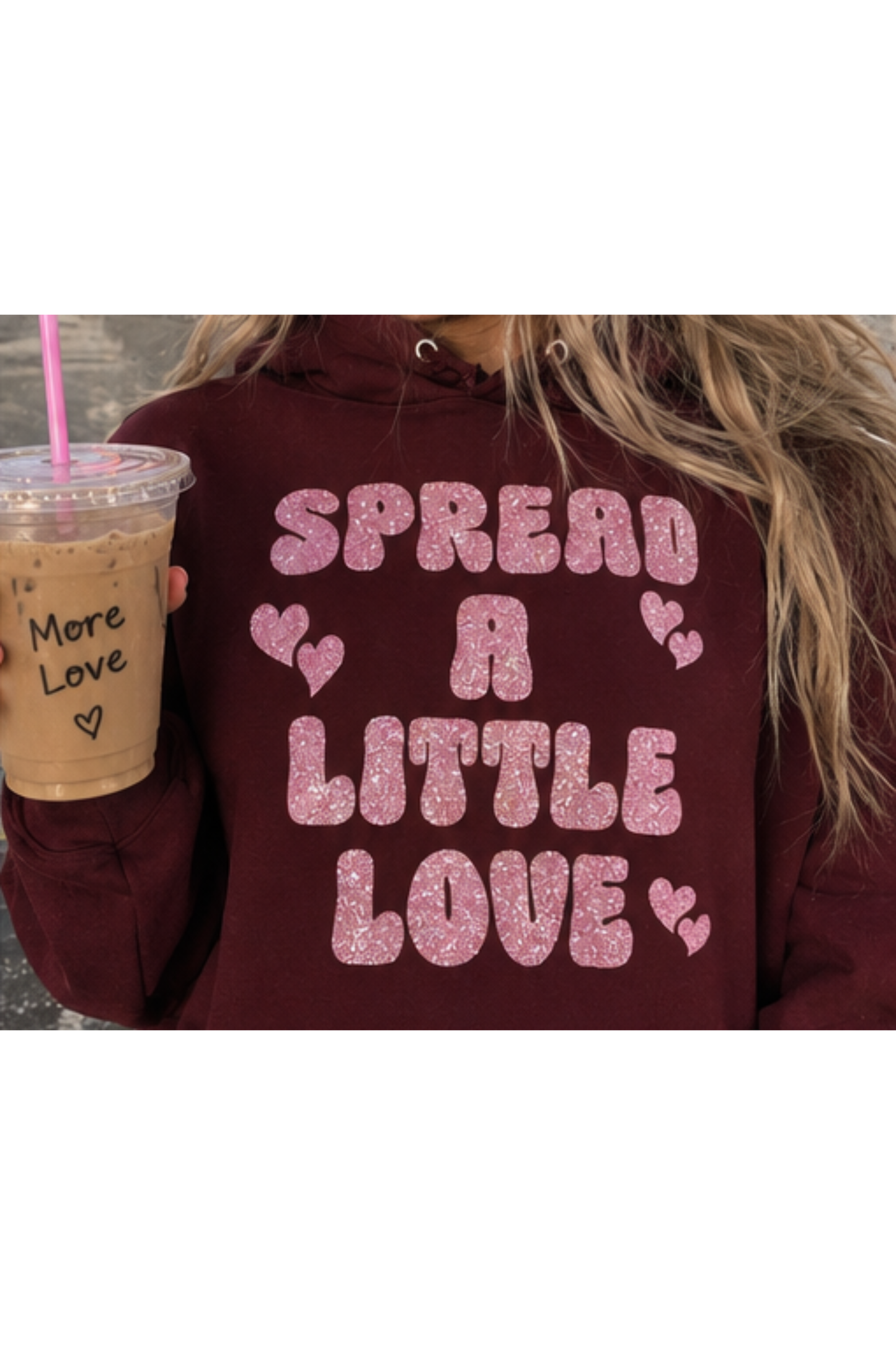 A person wearing a burgundy hoodie with pink glitter text that reads 'SPREAD A LITTLE LOVE' and small pink hearts, holding an iced coffee with a pink straw.