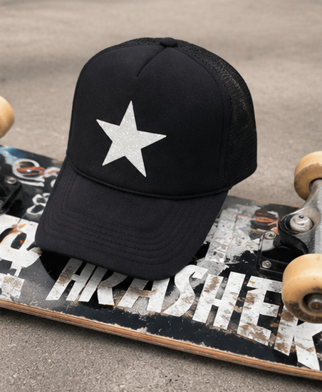 A black trucker-style baseball cap with a glittery silver star on the front, resting on a skateboard on a gray concrete surface.