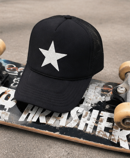 A black trucker-style baseball cap with a glittery silver star on the front, resting on a skateboard on a gray concrete surface.