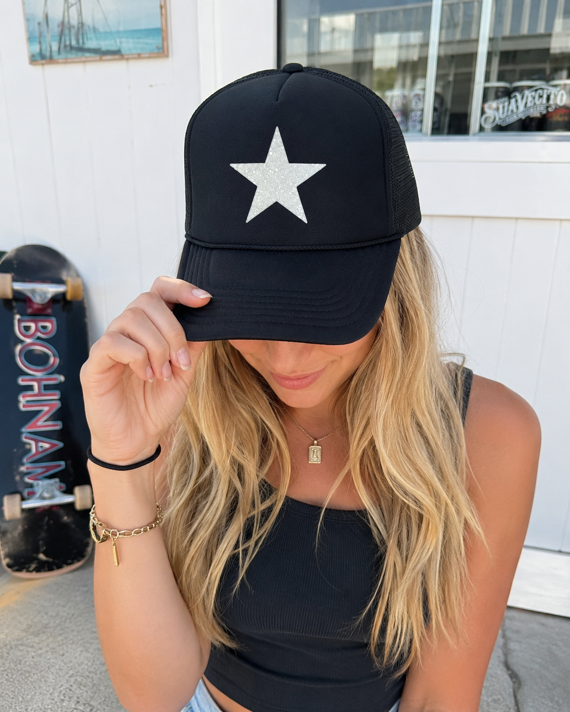 A woman wearing a black trucker-style baseball cap with a large white star on the front, holding the brim down over her eyes. She is wearing a black ribbed tank top and standing outdoors in front of a white building, with a skateboard visible to her side.