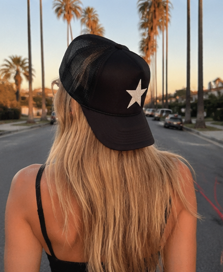 A woman viewed from behind wearing a black trucker-style baseball cap with a white star logo, standing on a palm-tree-lined street at sunset.