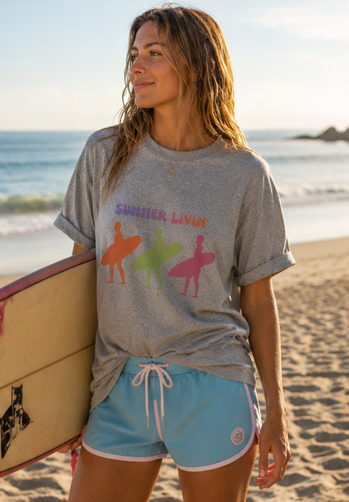 Woman on a beach wearing a gray t-shirt with colorful graphics and blue shorts, holding a surfboard.