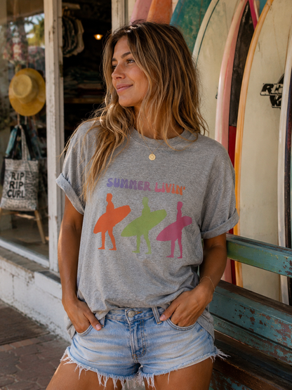 Woman wearing a gray t-shirt with colorful graphics and text, standing in front of surfboards.