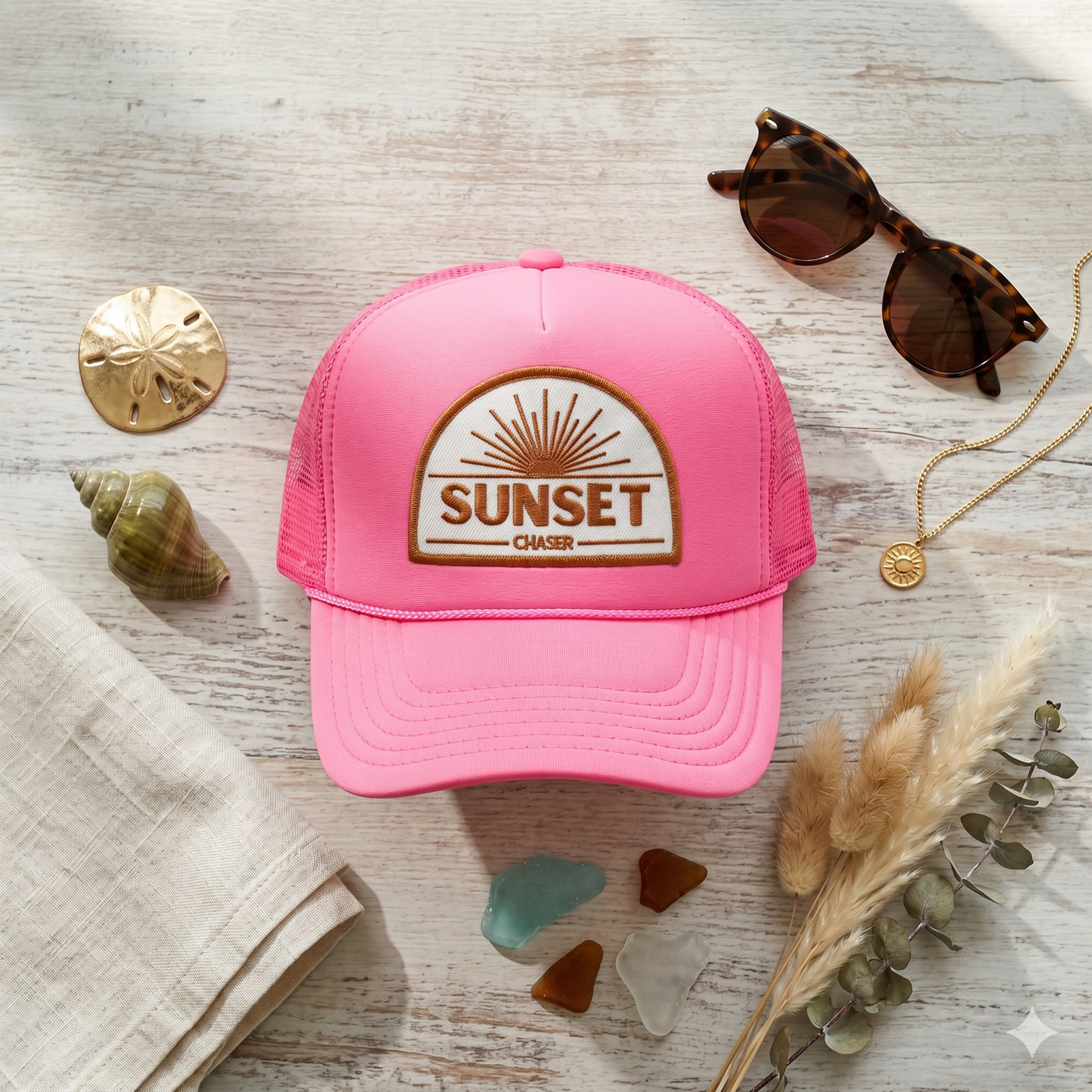 Pink cap with 'Sunset Chaser' logo on a wooden surface with sunglasses, shells, and dried plants.