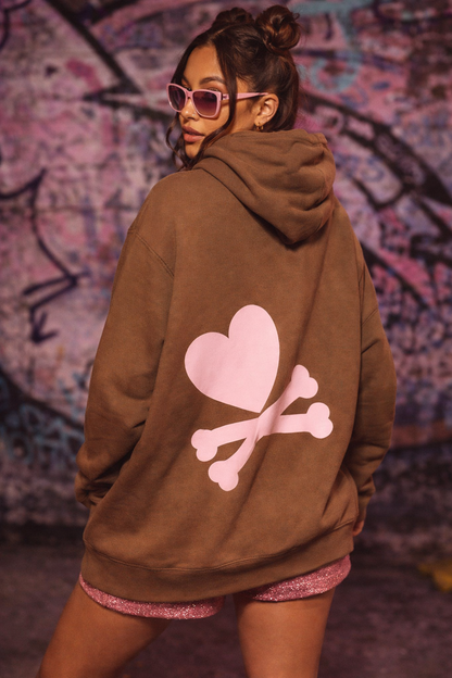 Person wearing a brown hoodie with a pink heart and bones design, standing against a graffiti-covered wall.