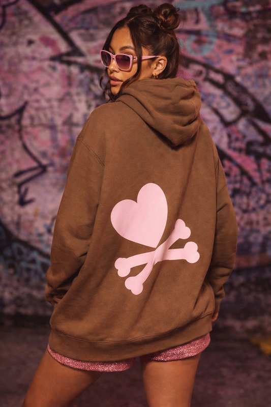Person wearing a brown hoodie with a pink heart and bones design, standing against a graffiti-covered wall.
