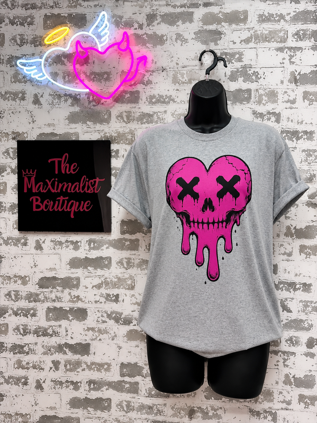 Gray t-shirt with pink skull heart design on a brick wall background, neon sign above.