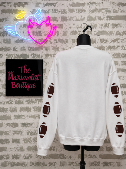 White sweatshirt with football designs on a mannequin against a brick wall with a neon sign above.