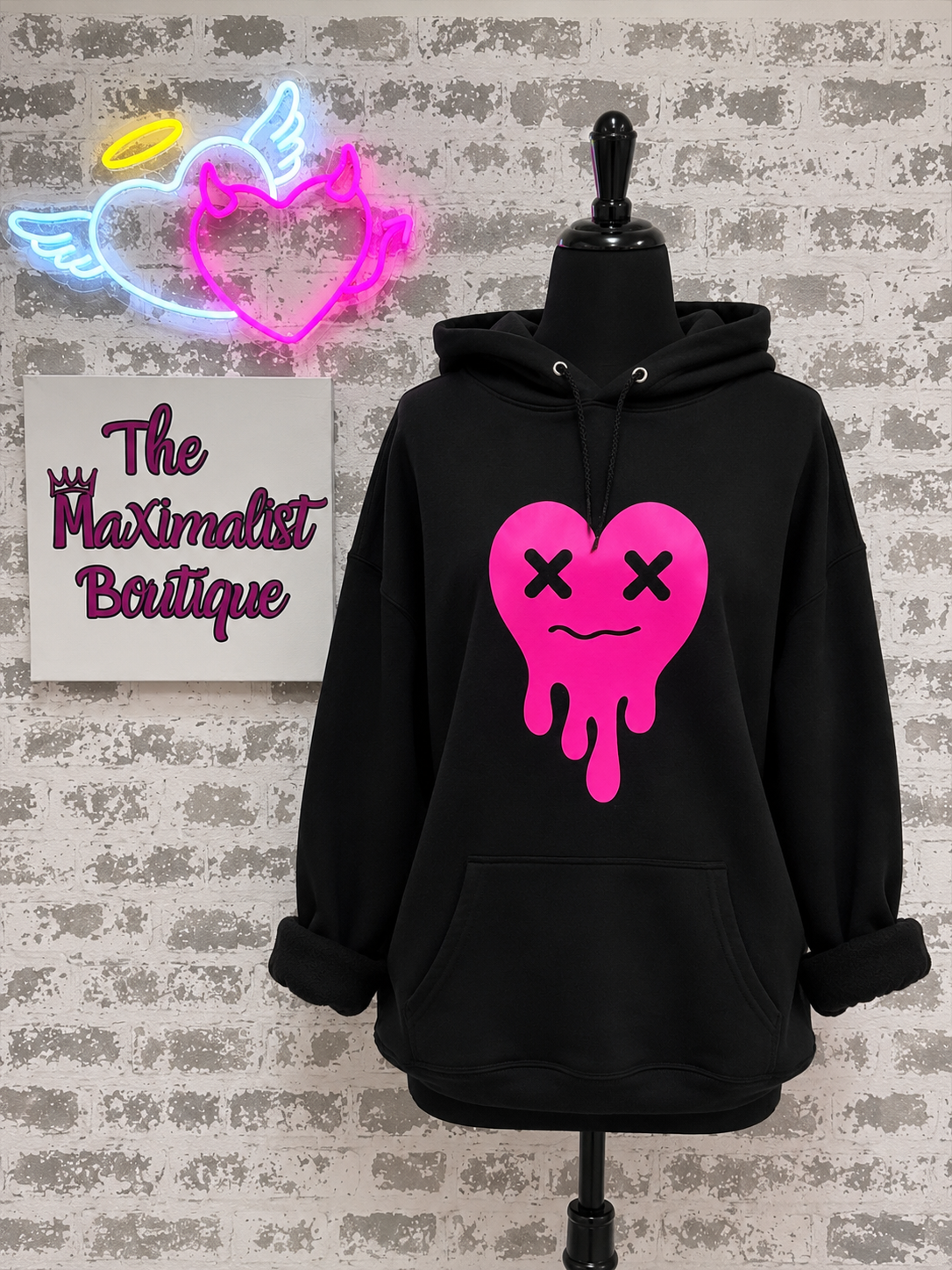A black pullover hoodie displayed on a black mannequin torso against a faux brick wall. The hoodie features a large bright pink dripping heart graphic on the front with black X eyes and a simple smile.