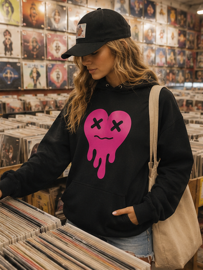 A woman in a record store wearing a black hoodie with a pink dripping heart graphic, a black baseball cap, and a beige shoulder bag, looking down at vinyl records on a shelf.