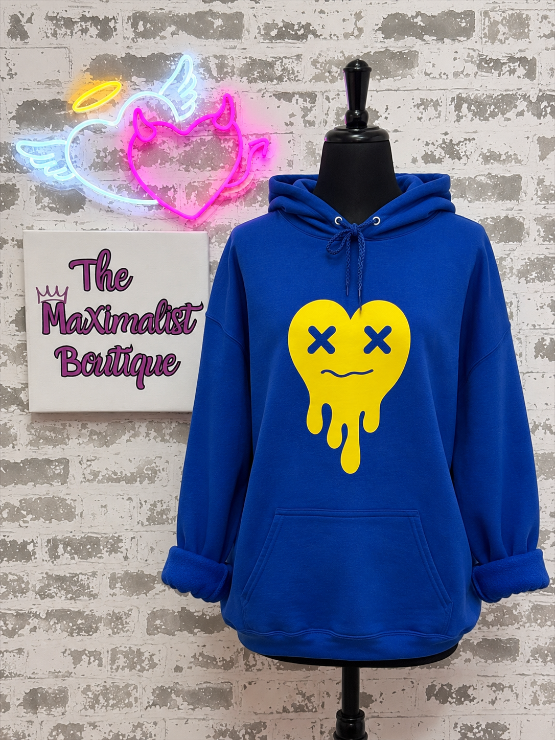 A royal blue hoodie displayed on a black mannequin torso against a faux brick wall. The hoodie features a large yellow dripping heart graphic with blue X eyes on the front.