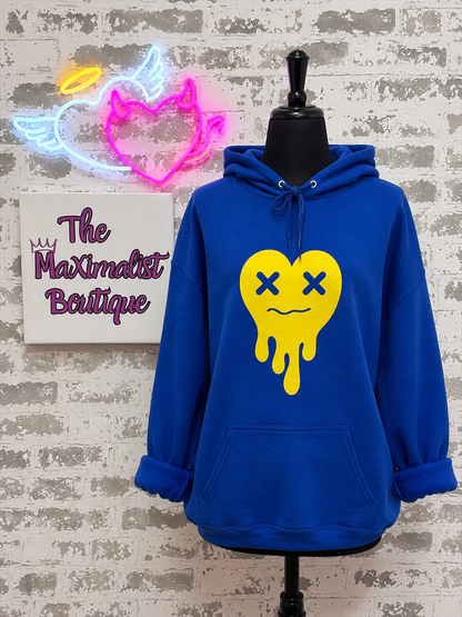 A royal blue hoodie displayed on a black mannequin torso against a faux brick wall. The hoodie features a large yellow dripping heart graphic with blue X eyes on the front.