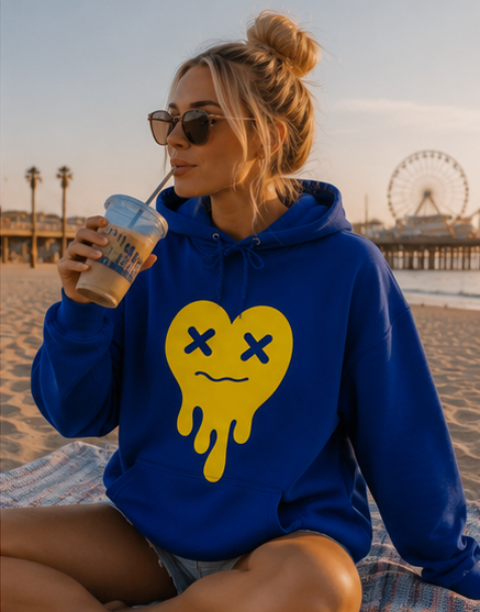 A woman sitting on a beach blanket at the beach, wearing a royal blue hoodie with a yellow dripping heart graphic with X eyes. She is holding an iced coffee.