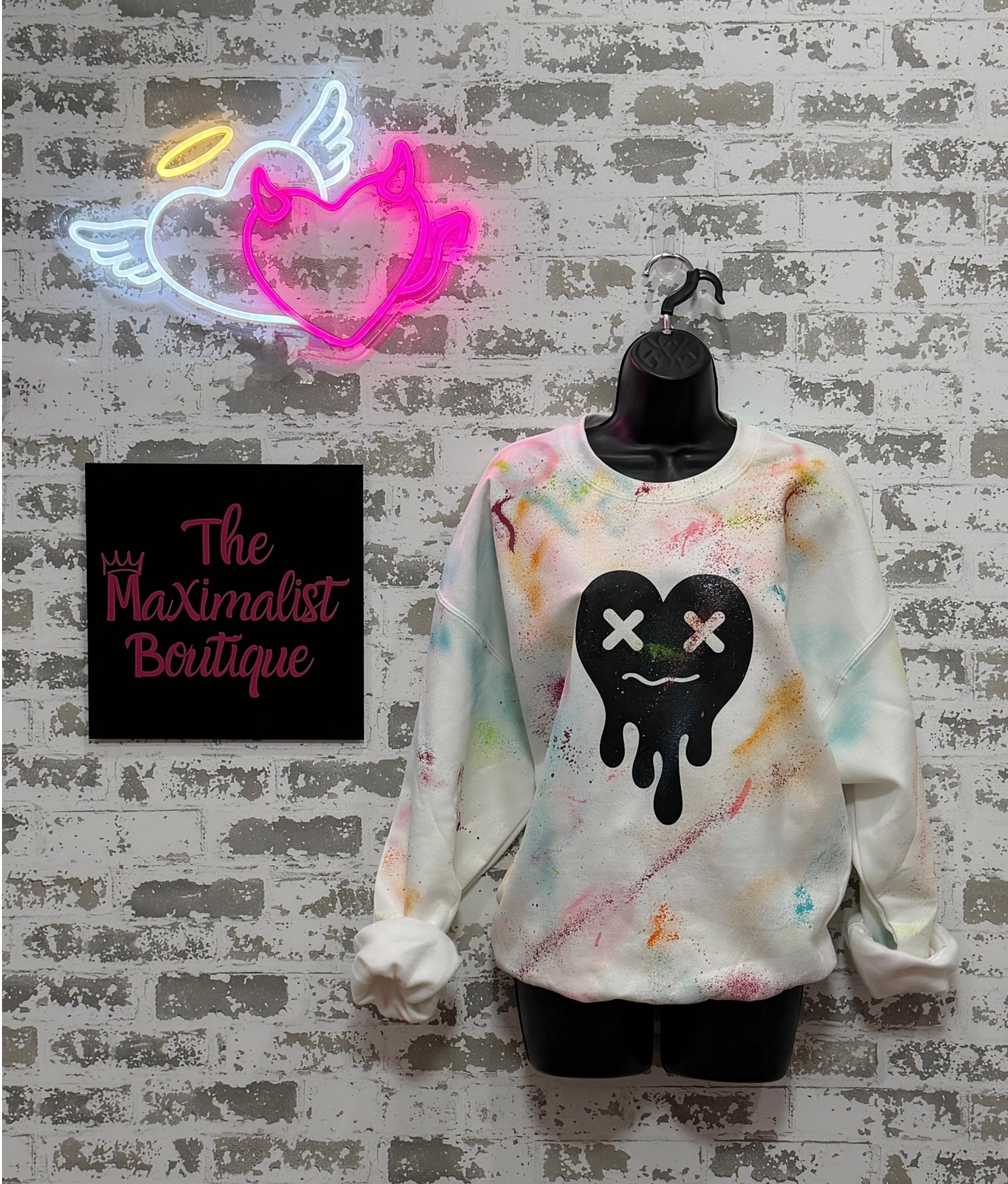Colorful handpainted sweatshirt with a black heart design on a hanger against a textured wall with 'The Maximalist Boutique' sign.