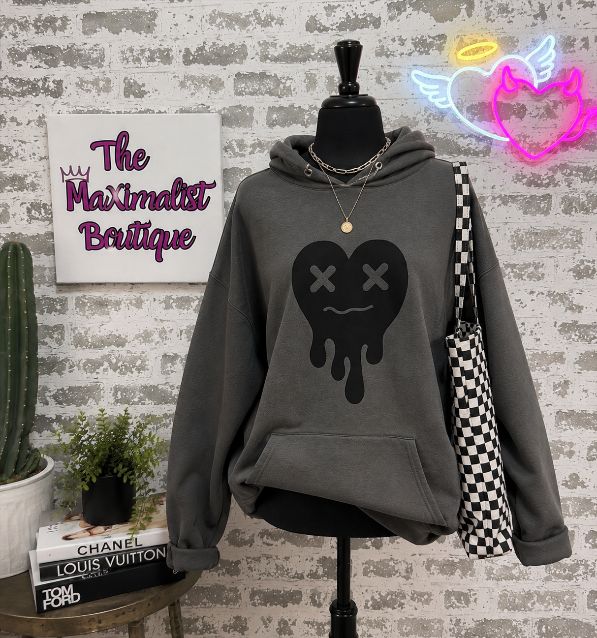 A dark gray hoodie displayed on a black mannequin torso against a faux brick wall. The hoodie features a black dripping heart graphic with X eyes on the front.