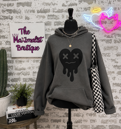 A dark gray hoodie displayed on a black mannequin torso against a faux brick wall. The hoodie features a black dripping heart graphic with X eyes on the front.