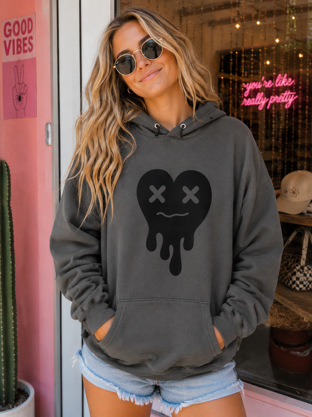 A woman wearing a dark gray hoodie with a black dripping heart graphic featuring X eyes, paired with denim shorts, standing in a boutique setting.