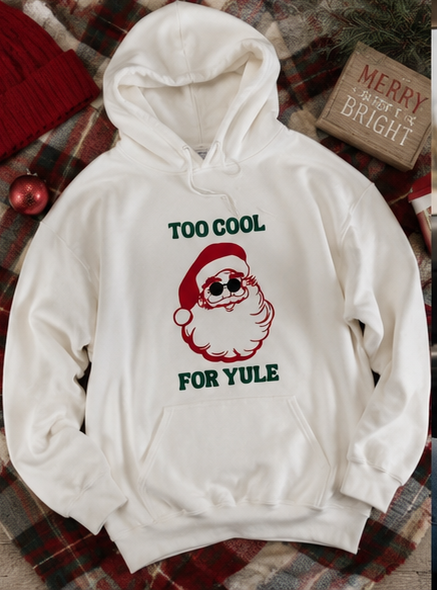 A white pullover hoodie with a Christmas graphic print featuring a Santa Claus illustration and the text 'TOO COOL FOR YULE' in green and red. The hoodie is laid flat on a red and green plaid blanket with holiday props, including a red knit sweater corner, a red ornament, and a wooden sign that reads 'MERRY AND THE BRIGHT'.