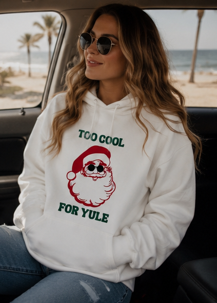 A woman wearing a white hoodie with a graphic of a Santa Claus wearing sunglasses and the text 'TOO COOL FOR YULE', sitting inside a car with a beach background visible through the window.