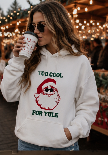 A woman wearing a white graphic hoodie with a red and green Santa Claus design and the text 'TOO COOL FOR YULE' holds a coffee cup in a festive outdoor market setting with warm string lights in the background.