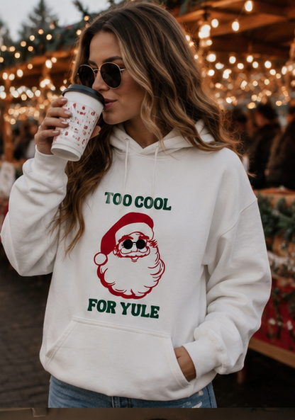 A woman wearing a white graphic hoodie with a red and green Santa Claus design and the text 'TOO COOL FOR YULE' holds a coffee cup in a festive outdoor market setting with warm string lights in the background.