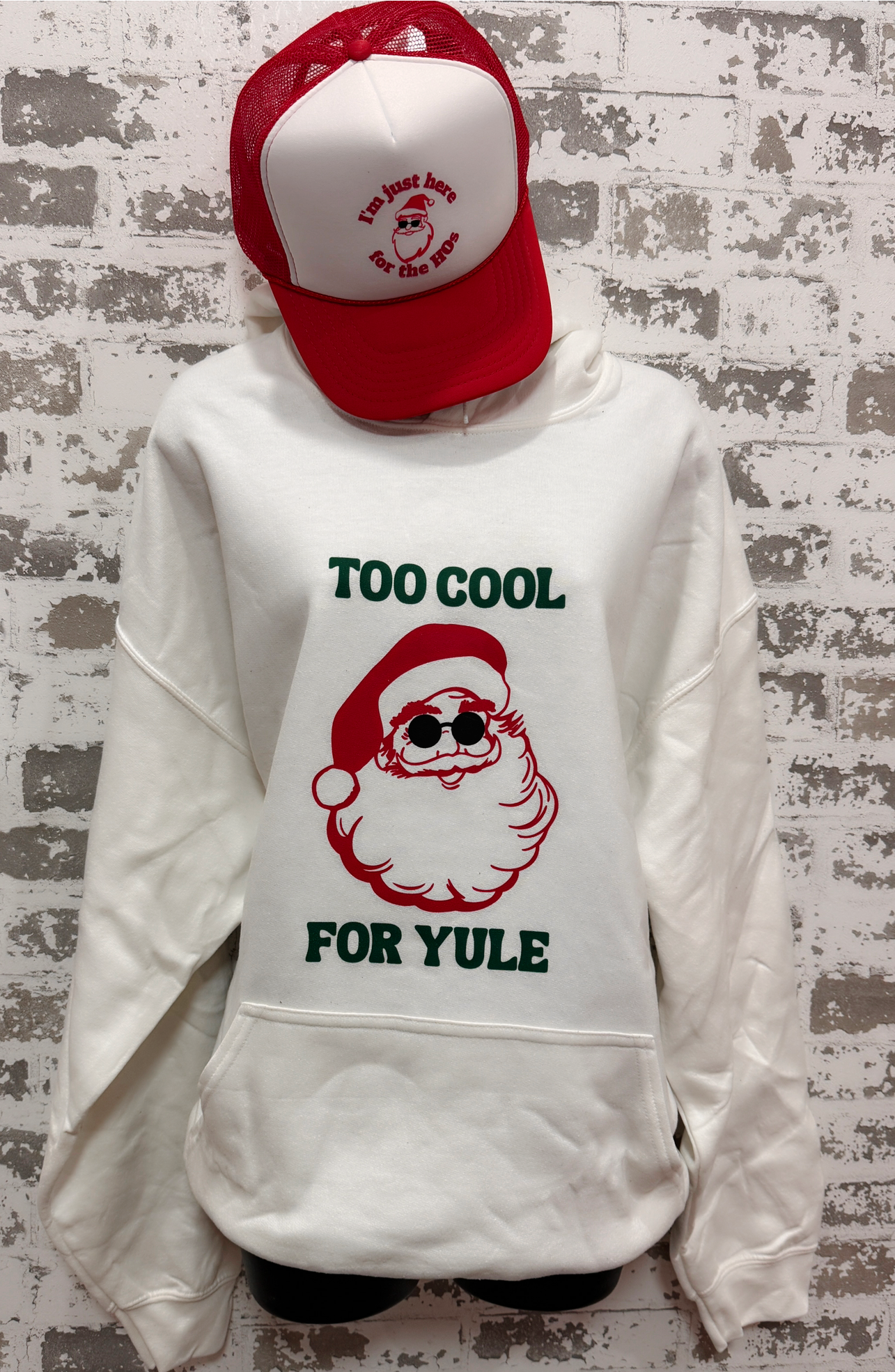 White sweatshirt with Santa Claus graphic and text 'Too Cool For Yule' worn by a person against a textured wall.