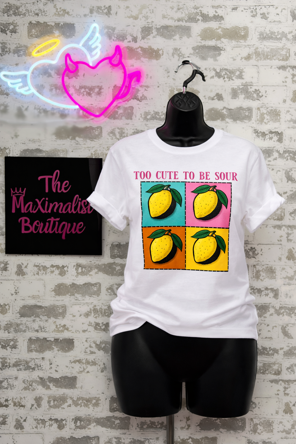 White t-shirt with lemon graphic and text on a mannequin against a brick wall with neon sign and 'The Maximalist Boutique' sign.