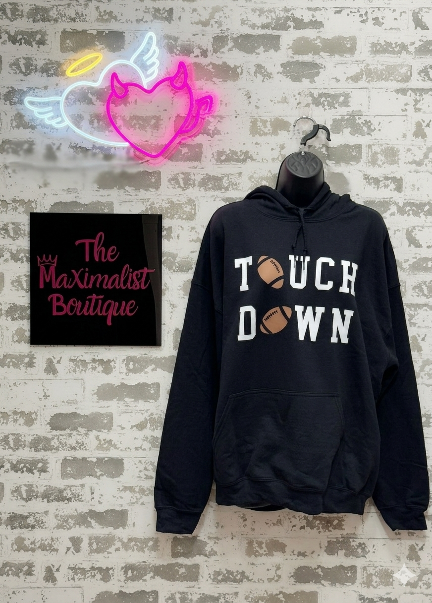 Black hoodie with 'Touchdown' text on a textured wall with neon sign and 'The Maximalist Boutique' sign.