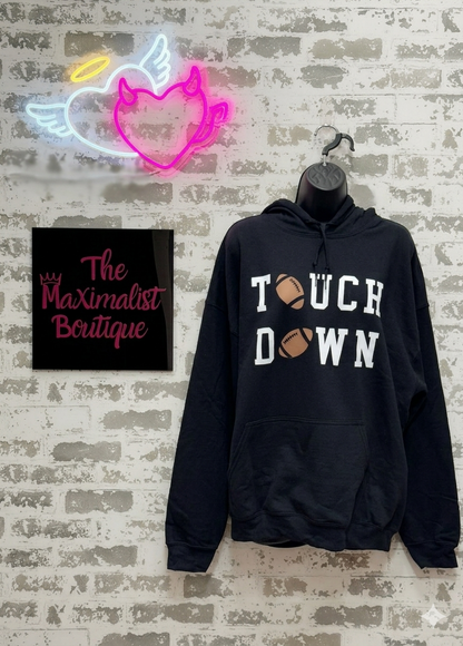 Black hoodie with 'Touchdown' text on a textured wall with neon sign and 'The Maximalist Boutique' sign.
