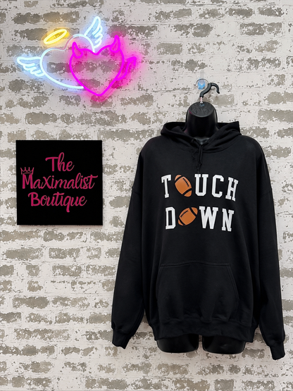 Black hoodie with 'Touchdown' text, neon signs, and 'The Maximalist Boutique' sign on a textured wall.