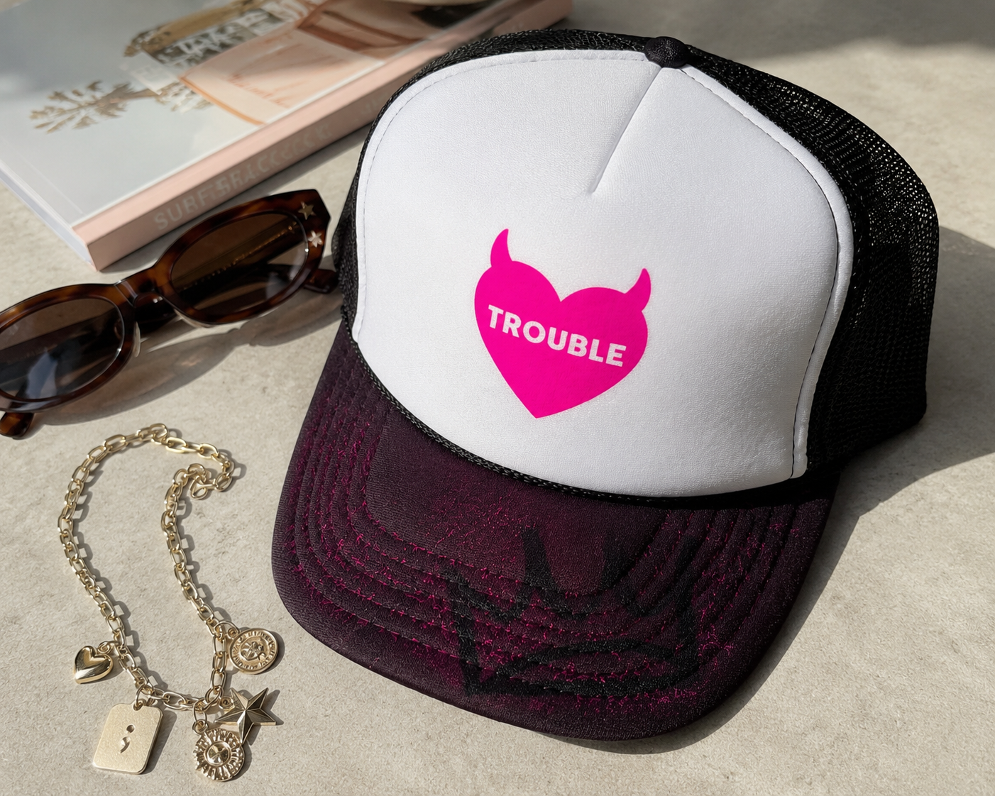 A white and black trucker hat with a pink devil-heart graphic that says 'TROUBLE' in white letters, styled on a light surface with sunglasses, a gold charm bracelet, and a book.
