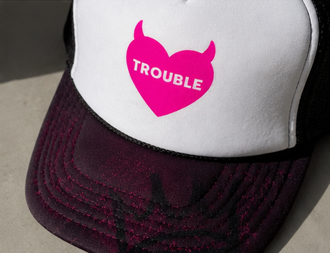 A close-up of a trucker hat featuring a white front panel with a pink devil-horn heart graphic containing the word 'TROUBLE' in white, and a dark purple brim with black mesh side panels.