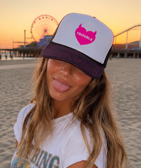 A woman on a beach at sunset wearing a white and dark purple trucker hat with a pink heart graphic that says 'TROUBLE'.