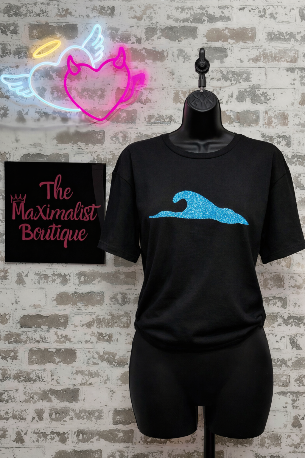 Black t-shirt with blue wave design on a mannequin against a brick wall with neon signs.