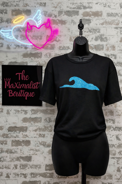 Black t-shirt with blue wave design on a mannequin against a brick wall with neon signs.