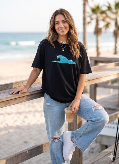 Woman wearing a black t-shirt with a blue graphic and ripped jeans, standing on a wooden deck by the beach.