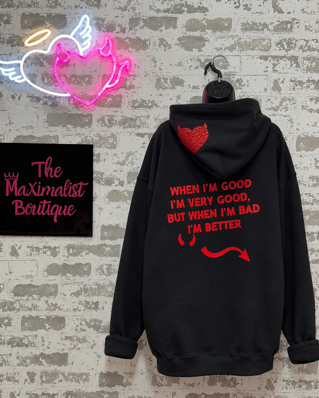 A black hoodie hanging on a hanger against a faux brick wall, featuring red text and graphic prints on the back. A neon heart sign and a 'The Maximalist Boutique' sign are visible in the background.