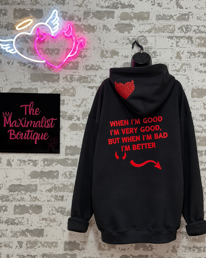 A black hoodie hanging on a hanger against a faux brick wall, featuring red text and graphic prints on the back. A neon heart sign and a 'The Maximalist Boutique' sign are visible in the background.