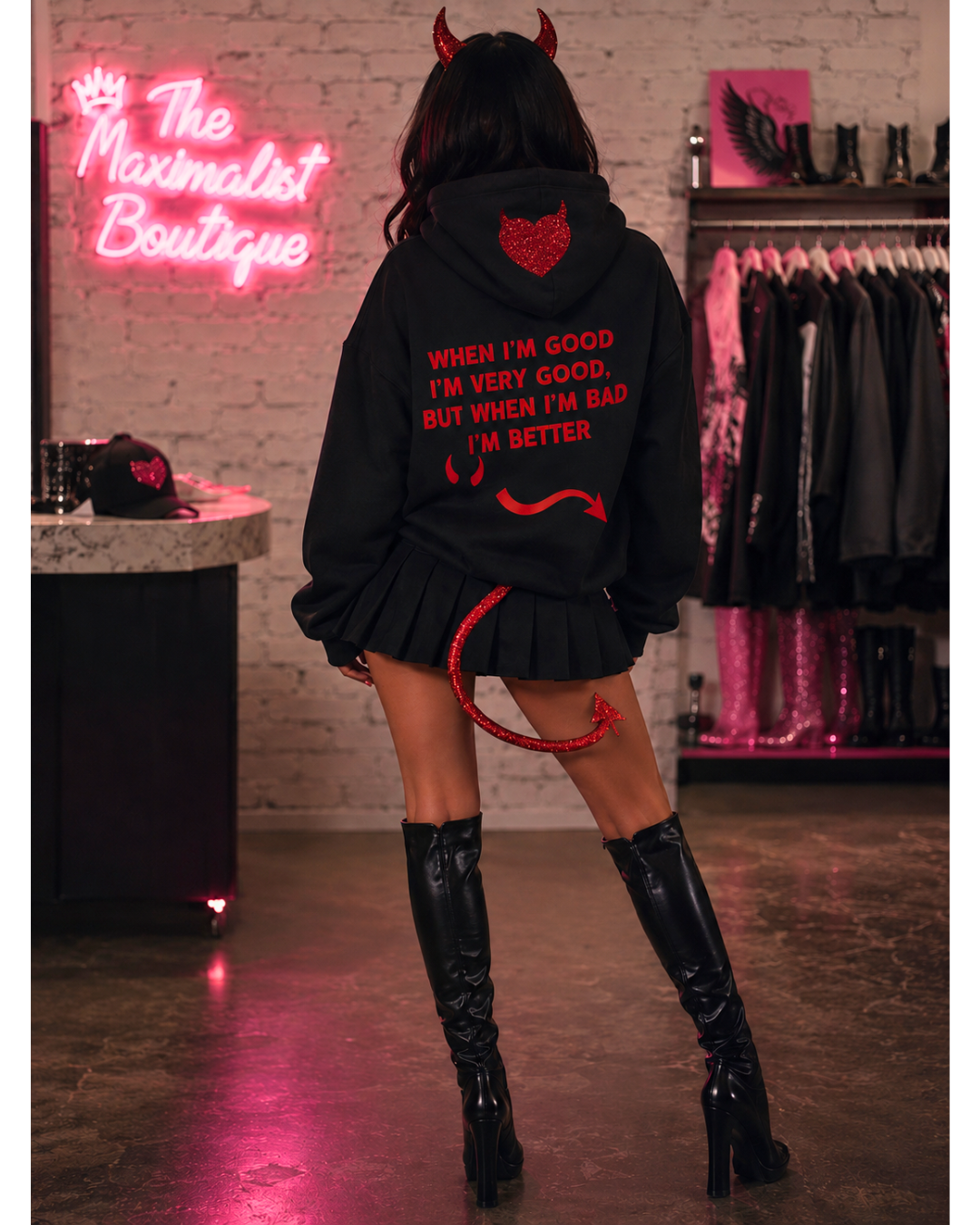 A model from behind wearing a black devil-themed hoodie with red text and graphics, styled with a pleated mini skirt and black knee-high boots in a boutique setting.