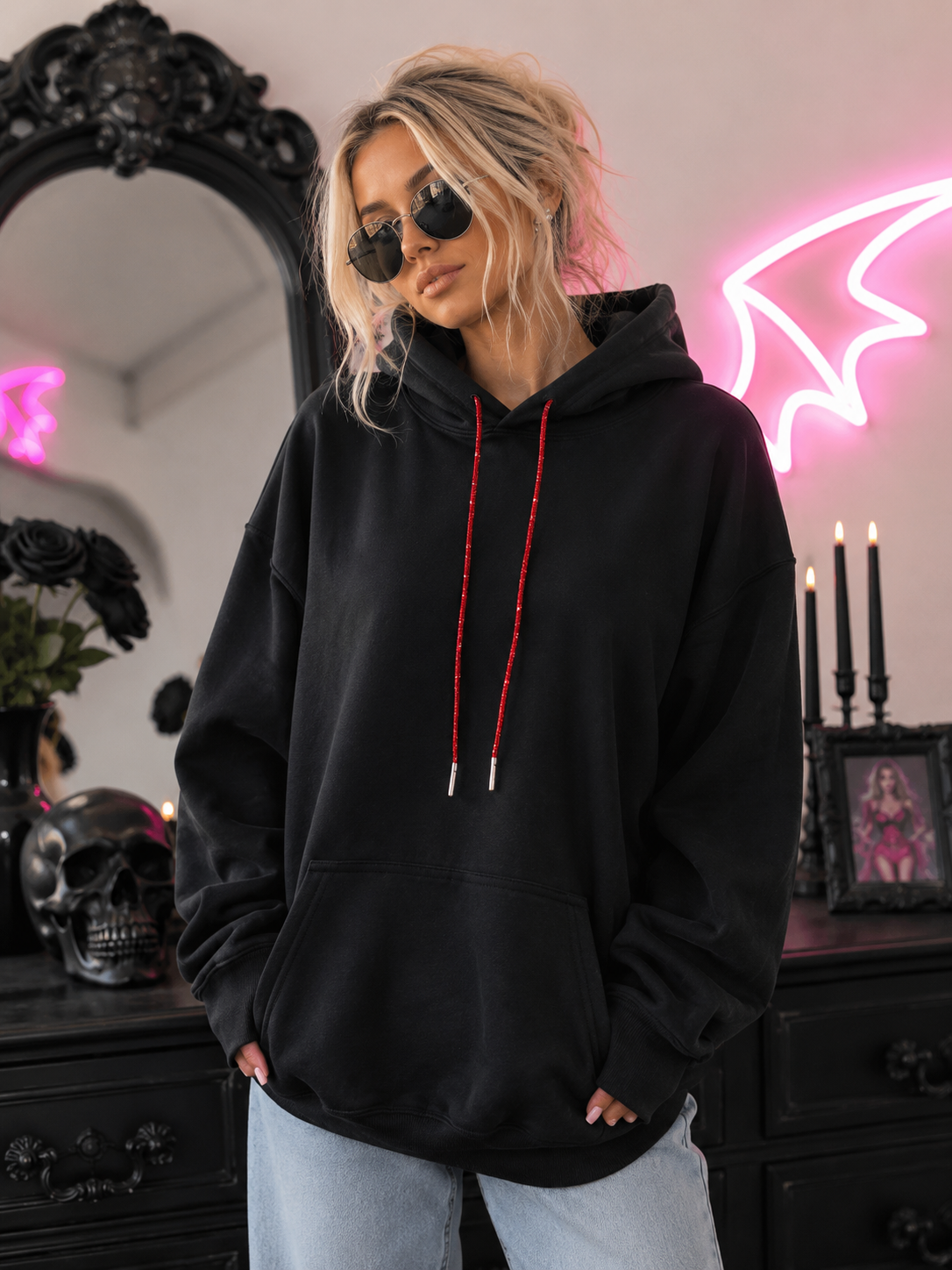 A woman wearing a black pullover hoodie with red drawstrings and light-wash jeans, standing in a gothic-themed room with a black mirror, skull decor, and pink neon lights.