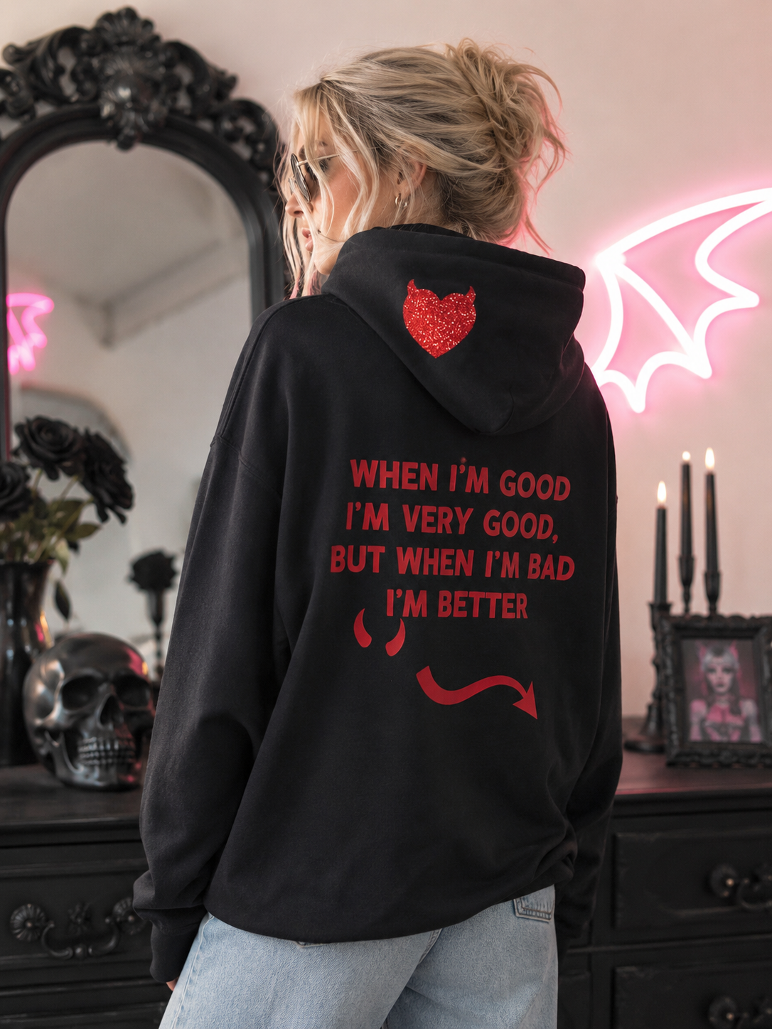 A woman shown from behind wearing a black hoodie with red devil-themed text and graphics, standing in a room with a mirror, candles, and a pink neon bat-wing light.
