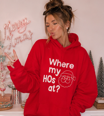A woman wearing a bright red hoodie with the white text 'Where my HOs at?' and a simple Santa outline graphic, posing in a decorated holiday room with a 'Merry Xmas' sign.
