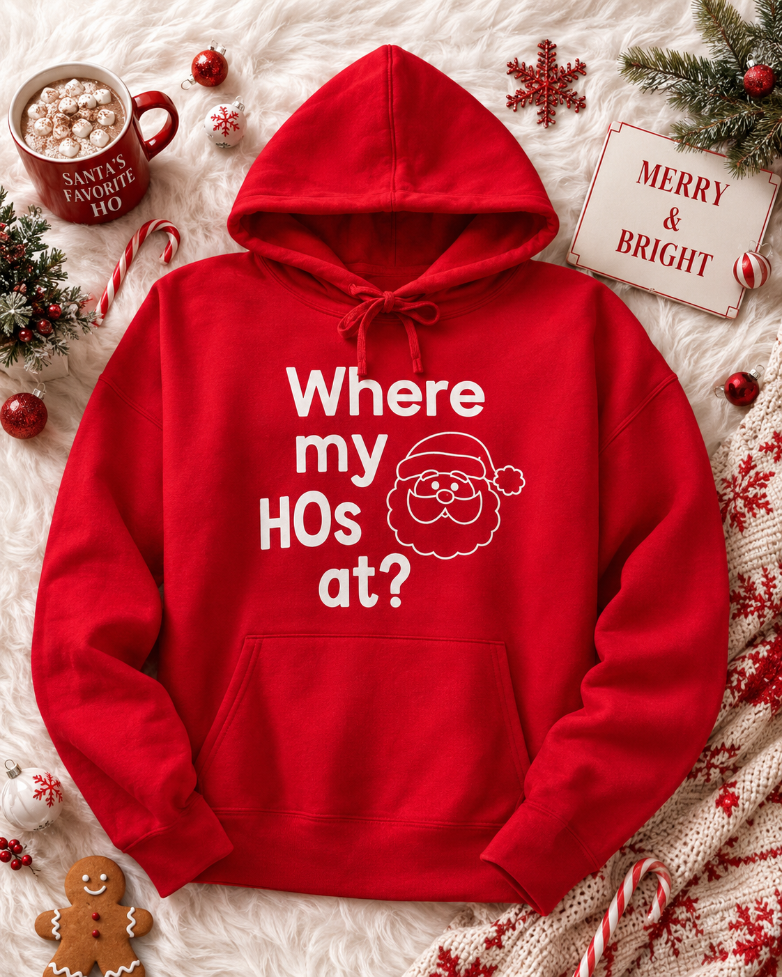 A red Christmas-themed hoodie with the white text 'Where my HOs at?' and a Santa face graphic, laid flat on a white faux fur background with holiday decorations including a mug of hot chocolate, candy canes, ornaments, a gingerbread cookie, and a 'Merry & Bright' sign.