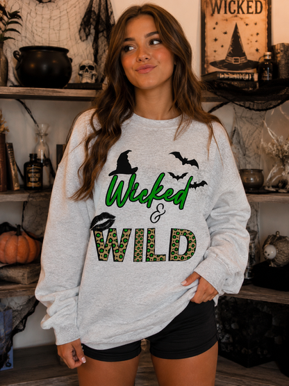 A woman wearing a light heather gray sweatshirt with a 'Wicked & Wild' Halloween graphic, standing in a decorated room with spooky props.