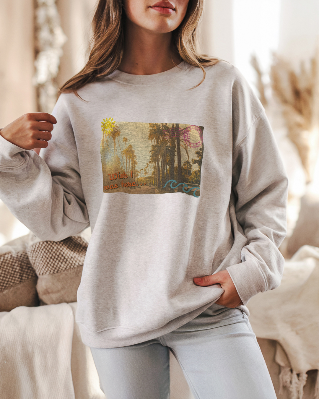 Gray sweatshirt featuring a vintage postcard-style palm tree scene with the phrase “wish I was here” in the bottom left corner on a model