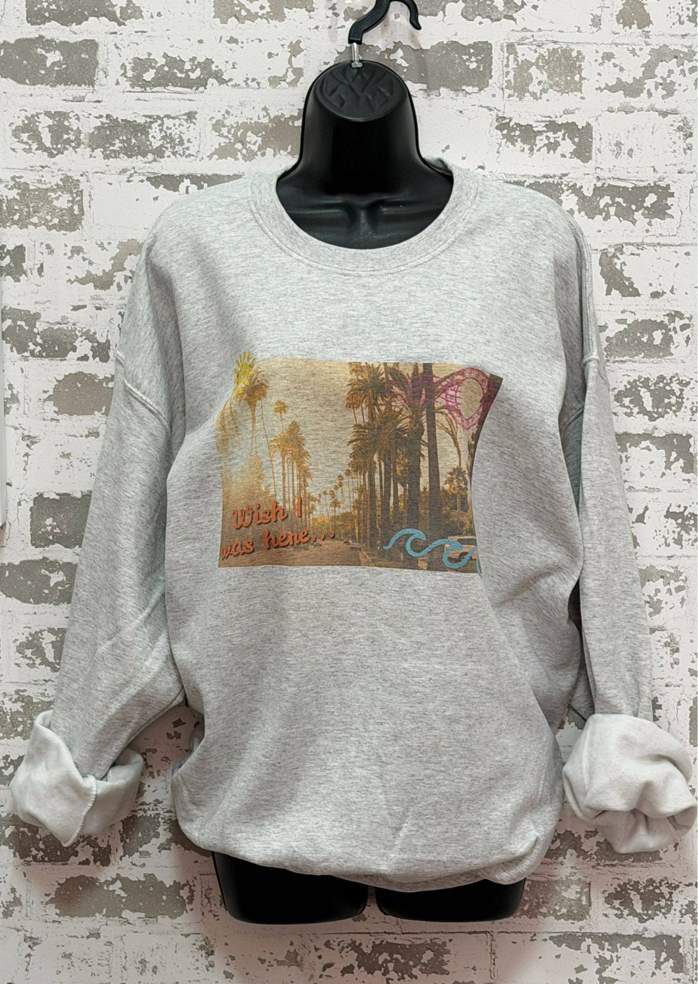 Gray sweatshirt featuring a vintage postcard-style palm tree scene with the phrase “wish I was here” in the bottom left corner